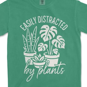 以下が含まれることがあります： 「EASILY DISTRACTED by plants」の白文字がプリントされたグリーンのTシャツ。スネークプラント、モンステラ、多肉植物など、さまざまな鉢植えの植物のイラストが描かれています。柔らかい素材のシャツです。