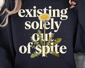 Op de afbeelding: Een marineblauwe sweatshirt met de tekst "existing solely out of spite" in een crèmekleurig lettertype. Een gele paardenbloem met groene bladeren staat centraal in het ontwerp. De sweatshirt is een casual, comfortabele stijl.