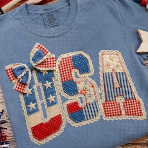 May include: Pale blue t-shirt featuring a patchwork design spelling "USA". The letters are made of various patterned fabrics, including stars, stripes, and floral prints, and are trimmed with lace. The shirt is decorated with a bow.