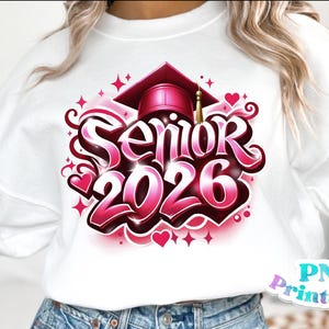 K&ouml;nnte beinhalten: Wei&szlig;es Sweatshirt mit einem pink-roten Aufdruck "Senior 2026". Das Design beinhaltet eine Abschlusskappe und Herzen. Das Sweatshirt ist ein l&auml;ssiges Kleidungsst&uuml;ck, passend f&uuml;r eine Abschlussfeier.