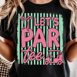 May include: Black t-shirt featuring the phrase "Let's Par Tee" in pink, with a golf cart graphic. The text is set against a mint green and black striped background. A pink bow is above the word "Par".