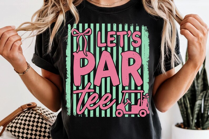 May include: Black t-shirt featuring the phrase "Let's Par Tee" in pink, with a golf cart graphic. The text is set against a mint green and black striped background. A pink bow is above the word "Par".