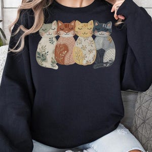 Pu&ograve; includere: Una felpa blu navy con un design ricamato di quattro gatti stilizzati. Ogni gatto ha un motivo e un colore unici, tra cui verde, marrone, giallo e grigio. I gatti sono disposti uno accanto all'altro.
