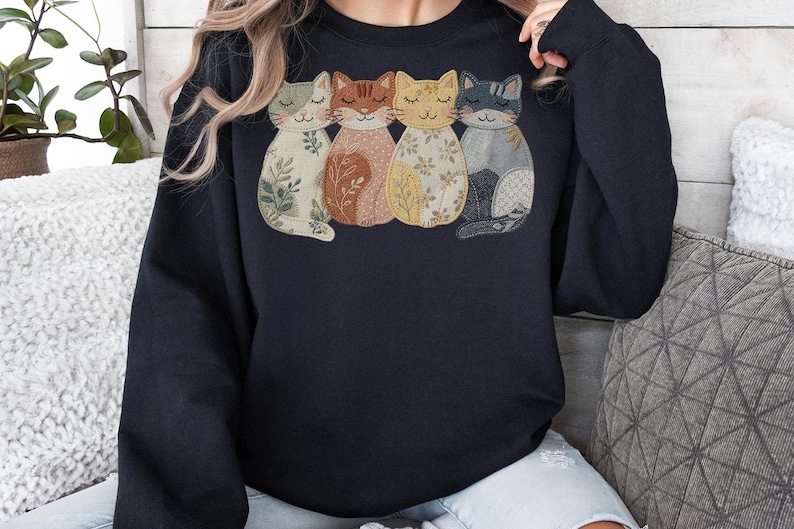 Pu&ograve; includere: Una felpa blu navy con un design ricamato di quattro gatti stilizzati. Ogni gatto ha un motivo e un colore unici, tra cui verde, marrone, giallo e grigio. I gatti sono disposti uno accanto all'altro.