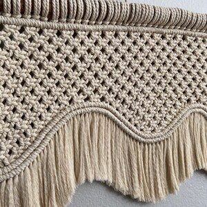 May include: A beige macrame wall hanging with a scalloped bottom and fringe. The hanging is made with natural cotton cord and has a wooden dowel at the top.