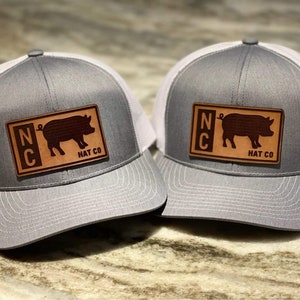 以下が含まれることがあります： グレーと白のトラッカーハット2つ。茶色の革のパッチには「NC」の文字と豚のシルエットが描かれている。パッチには「Hat Co.」の文字も書かれている