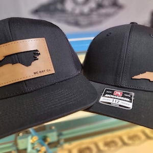 Może przedstawiać: Dwie czarne czapki typu trucker z brązową skórzaną naszywką z konturem stanu Karolina Północna. Jedna czapka ma napis "NC HAT Co." na naszywce. Druga ma metkę Richardson 112 Youth.