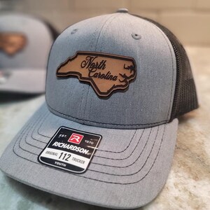 Może przedstawiać: Szara i czarna młodzieżowa czapka trucker z brązową skórzaną naszywką przedstawiającą zarys Karoliny Północnej i napis "North Carolina". Czapka jest wykonana przez Richardson i ma napis "Original 112 Trucker Youth" z przodu.