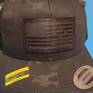 Könnte beinhalten: Eine schwarze und grüne Tarnkappe mit einem schwarzen Lederpatch, auf dem "2nd Amendment Since 1791" steht. Der Patch zeigt eine stilisierte amerikanische Flagge mit gekreuzten Gewehren.