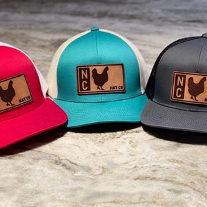 Può includere: Tre cappellini da camionista con rete bianca sul retro e una toppa in pelle marrone con una silhouette di pollo e il testo "NC HAT CO". I cappellini sono rosso, turchese e grigio.