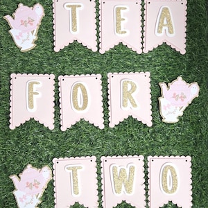 May include: A pink banner with the words "TEA FOR TWO" in gold glitter letters. The banner has a scalloped edge and is decorated with teapot cutouts. The banner is set against a green background.