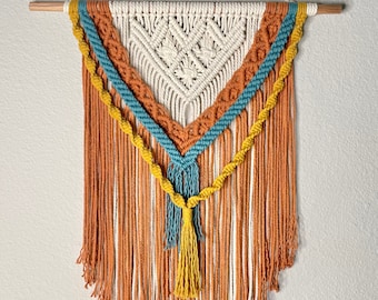 Pode incluir: Uma tapeçaria de macramé triangular com um centro branco e franjas laranja, azul e amarelo. As franjas são feitas de fio e têm uma borla na parte inferior.
