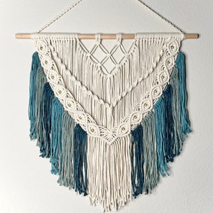 Può includere: Un arazzo da parete in macramè bianco con un motivo geometrico e frange blu e verdi. L'arazzo è decorato con perline dorate.