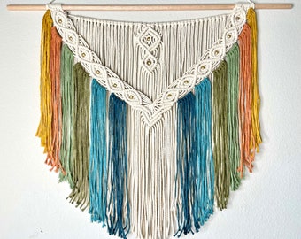 Pode incluir: Um pendurador de parede em macramé com uma barra de madeira. O design apresenta uma seção central creme com um padrão geométrico e detalhes dourados. Franjas coloridas em tons de amarelo, laranja, verde e azul adicionam um toque vibrante a esta decoração boêmia.