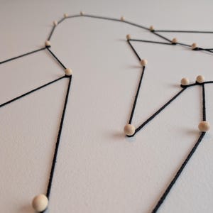 Può includere: Un'opera d'arte da parete composta da un sottile filo nero teso tra piccole perline di legno di colore chiaro. Il filo forma un motivo geometrico e lineare su una superficie piana, creando un design simile a una costellazione con punti interconnessi.