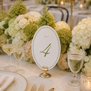 Può includere: Un'elegante mise en place con un centrotavola floreale e il numero del tavolo 4. Il segnaposto ovale ha un bordo e un testo verdi. Piatti, posate e bicchieri con bordi dorati completano l'allestimento.
