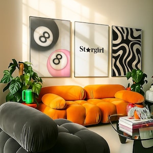 May include: An interior design scene featuring an orange modular sofa, a dark gray sofa, and a glass coffee table. Artwork includes a black and white abstract print, a "Star Girl" print, and a pool ball print. Green plants and decorative items complete the space.