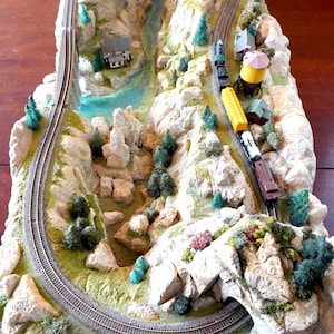 May include: A model train set featuring a winding track, a trestle bridge, and a train with yellow and black carriages travelling through a mountainous landscape with rock formations and trees.