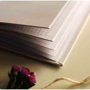 May include: A closed notebook with a grid pattern on the pages, a small bouquet of purple flowers, and a piece of twine on a beige surface.