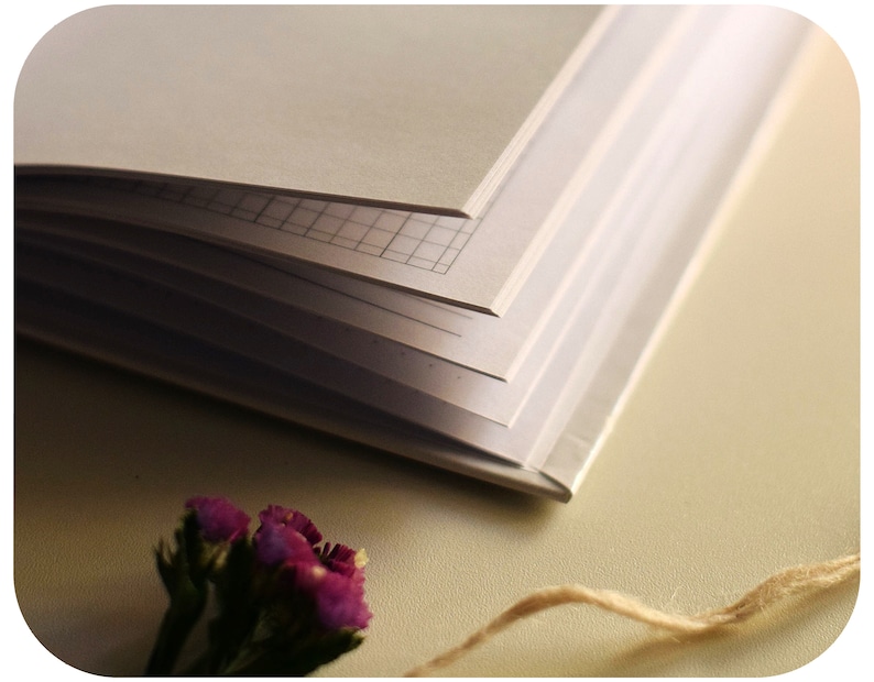 May include: A closed notebook with a grid pattern on the pages, a small bouquet of purple flowers, and a piece of twine on a beige surface.