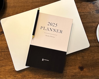 May include: A black and cream planner with the text "2025 PLANNER" and "Sarah O'Brien" on the cover. The planner is open to a black page with the word "HABITONIA" printed on it.