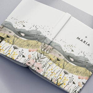Può includere: Un quaderno bianco con un'illustrazione ad acquerello di un paesaggio con colline, fiori e foglie. Il quaderno ha il testo "The Habito" sulla copertina anteriore e la parola "MARIA" sulla copertina posteriore.