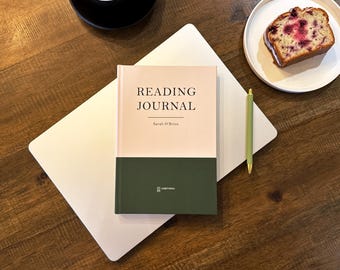 May include: A reading journal with the title "Reading Journal" and the author's name, Sarah O'Brien, is on a white laptop. A green pen and a slice of cake on a white plate are also visible. The journal has a two-tone cover.