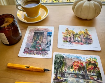 Op de afbeelding: Een stilleven met aquarelpostkaarten met bezienswaardigheden van Boston, een gele koffiekop en schotel, een kleine witte pompoen, een kaars en een gele vulpen. De ansichtkaarten tonen Beacon Hill, Harvard Yard en de Boston Public Garden.