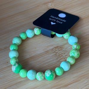 May include: A beaded bracelet featuring a variety of green and turquoise-colored stones. The bracelet is strung on a clear elastic cord. A black tag with the words "Handmade with love" is attached to the bracelet.