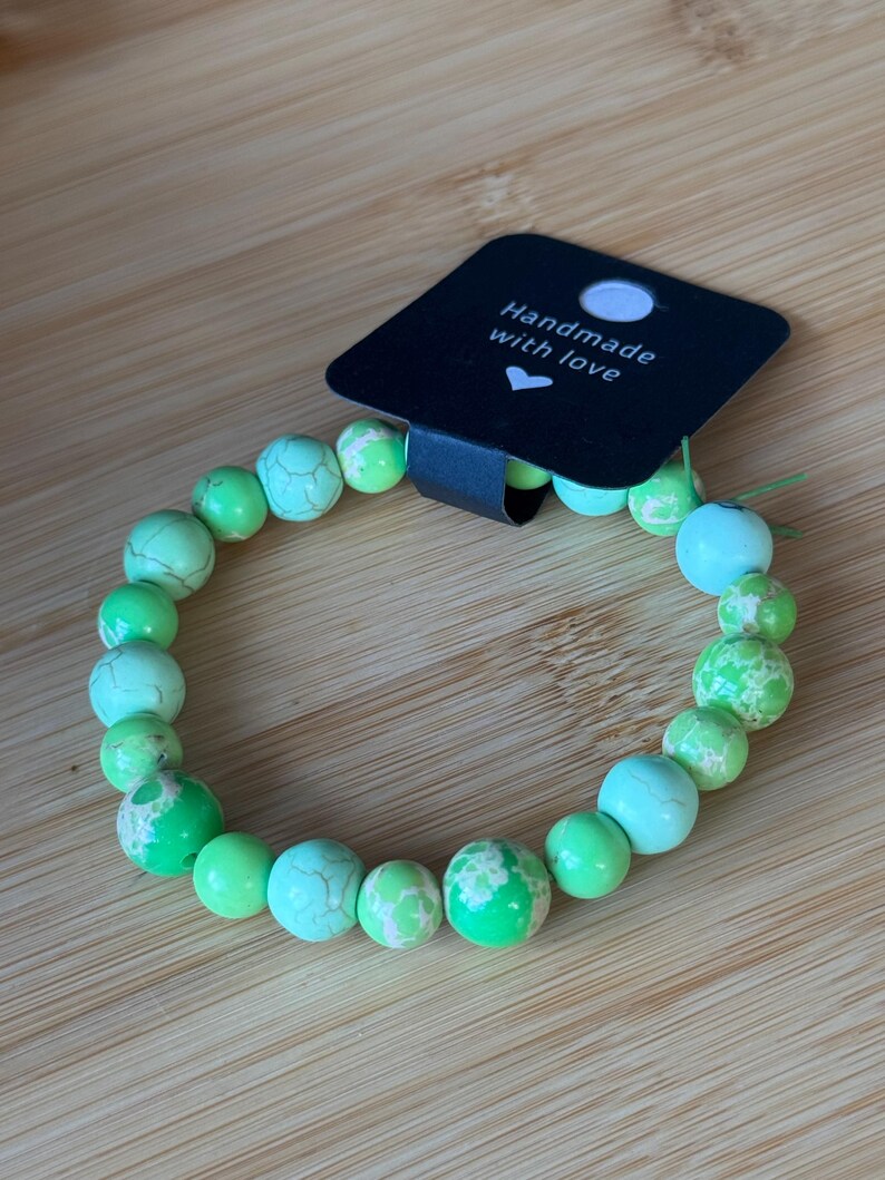May include: A beaded bracelet featuring a variety of green and turquoise-colored stones. The bracelet is strung on a clear elastic cord. A black tag with the words "Handmade with love" is attached to the bracelet.