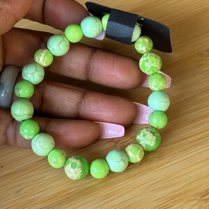 May include: A beaded bracelet featuring a mix of green and light green round beads. Some beads have a textured, cracked appearance. The bracelet is held against a wooden surface.