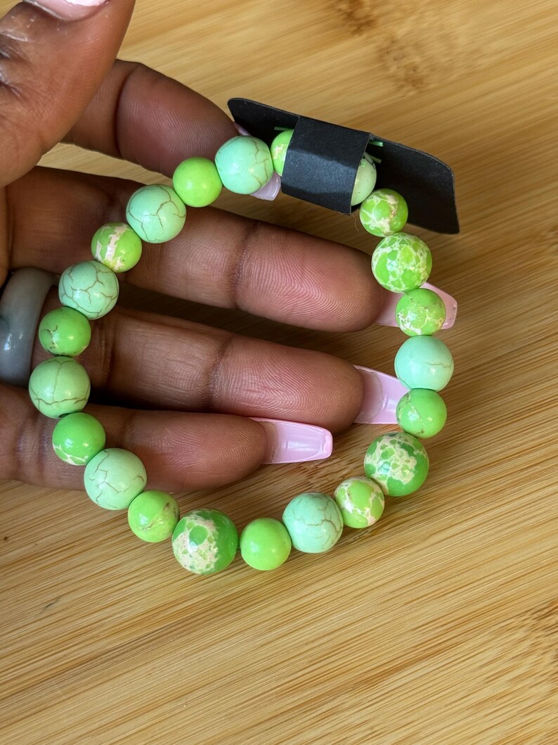 May include: A beaded bracelet featuring a mix of green and light green round beads. Some beads have a textured, cracked appearance. The bracelet is held against a wooden surface.