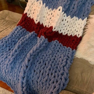 May include: A chunky knit throw blanket with stripes of blue, white, and burgundy. The blanket is made of soft, fluffy yarn and is perfect for cuddling up on the couch.