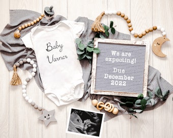 Peut inclure: Une grenouillère blanche avec le texte "Baby Varner" dessus, une guirlande de perles grises et blanches, une étoile en bois, une lune en bois en forme de croissant, un tableau à lettres avec le texte "We are expecting! Due December 2022", un anneau en bois, un petit cadre blanc avec une image d'échographie et des branches vertes feuillues sur un fond de bois clair.
