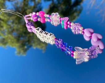 May include: A purple and pink beaded phone charm with a butterfly charm, a smiley face bead, and the word "ARMY" spelled out in white letter beads.