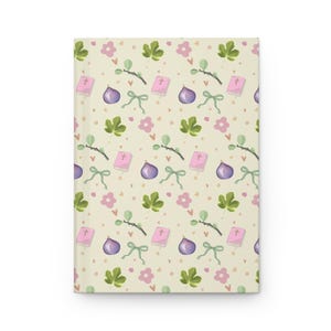 Könnte beinhalten: Ein geschlossenes Hardcover-Journal mit einem sich wiederholenden Muster aus Feigen, rosa Blumen, grünen Blättern, Schleifen und rosa Büchern mit Kreuzen auf cremefarbenem Hintergrund. Das Journal hat ein mattes Finish.