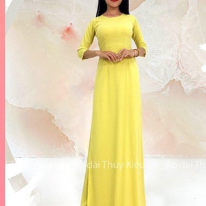 May include: A yellow maxi dress with a round neckline and three-quarter sleeves. The dress has a simple design and is made of a lightweight fabric. The model is wearing the dress and is standing in front of a white background. The text "MÃ: AK25" is visible at the bottom of the image.