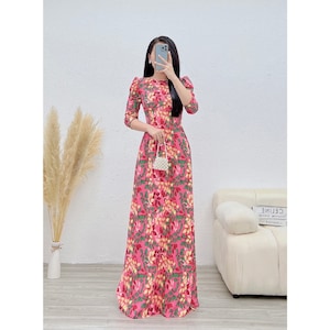 Könnte beinhalten: Ein pinkfarbenes Maxikleid mit einem Blumendruck mit grünen Blättern, gelben Blüten und rosa Akzenten. Das Kleid hat einen runden Ausschnitt und 3/4 Ärmel. Das Model hält eine kleine weiße Perlenhandtasche.