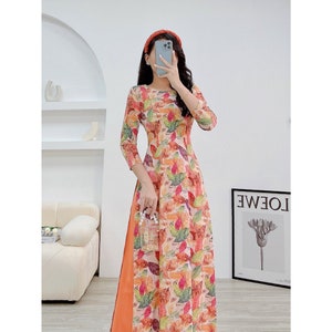 Op de afbeelding: Een lange, vloeiende jurk met een bloemenprint met bladeren en bloemen in tinten oranje, roze en groen. De jurk heeft een getailleerd bovenlijf en een vloeiende rok. De jurk is versierd met een perzikkleurige band.