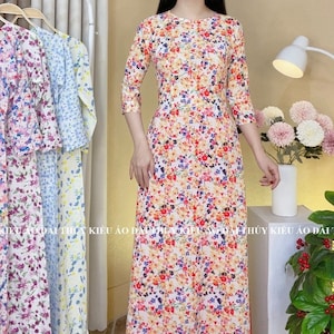 May include: A white long-sleeved dress with a floral print in shades of red, orange, yellow, and blue. The dress has a round neckline and three-quarter sleeves. The dress is shown on a model standing in front of a white background.