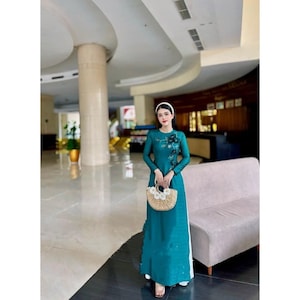 May include: A woman wearing a teal blue dress with floral embroidery and a white headband. She is holding a woven straw bag with white flowers.