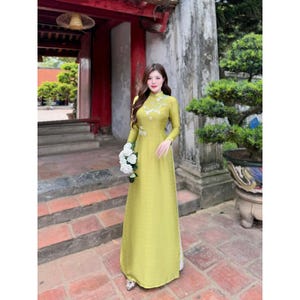 May include: A woman wearing a long, light green dress with white floral embroidery. She is holding a bouquet of white flowers and standing in front of a traditional building.