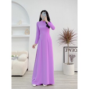 Könnte beinhalten: Ein langes, lavendelfarbenes Kleid mit einem Mandarinkragen und langen Ärmeln. Das Kleid hat einen taillierten Oberteil und einen fließenden Rock.