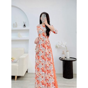 Könnte beinhalten: Ein weißes Maxikleid mit orangefarbenem Blumendruck und einem Schlitz an der Seite. Das Kleid hat einen runden Ausschnitt und Dreiviertelärmel.