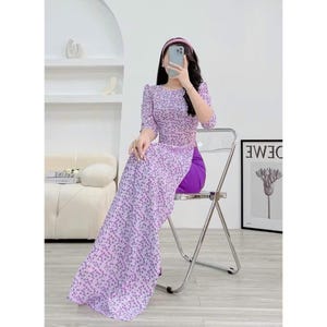 Puede incluir: Una mujer con un vestido largo de flores blancas y moradas con una falda morada. Está sentada en una silla de plástico transparente.