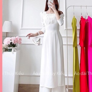 May include: A white long-sleeved dress with a square neckline and a pleated bodice. The dress has a fitted waist and a flowing skirt. The dress is paired with white wide-leg trousers. The model is holding a white handbag with pearl accents. The text "ΜΑ: A13" is visible at the bottom of the image.