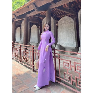 Puede incluir: Una mujer con un vestido largo de manga larga de color lavanda con bordados florales. El vestido tiene un cuello alto y una abertura lateral. Está de pie frente a una pared de piedra con una estructura de madera detrás de ella.