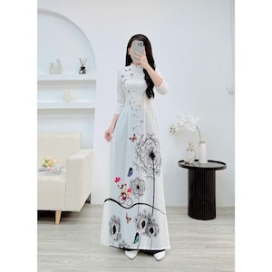 Puede incluir: Un vestido blanco con estampado de diente de león y mariposa. El vestido tiene cuello mandarín y mangas largas. El vestido es de largo hasta el suelo y tiene un corpiño ajustado.
