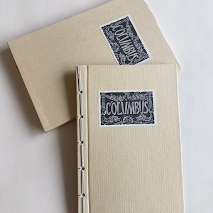 Pode incluir: Um diário creme e sua caixa correspondente. O diário é encadernado à mão com costuras visíveis ao longo da lombada. Tanto o diário quanto a caixa têm um rótulo preto e branco que diz "COLUMBUS" em uma fonte ornamentada.