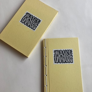 Pode incluir: Um conjunto de diário e caixa amarelo claro. O diário é costurado à mão com fio prateado. Tanto o diário quanto a caixa têm um adesivo em preto e branco que diz "MASKS UNMASKS". O conjunto está em um fundo branco.